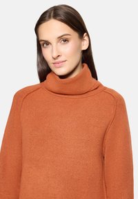 Femme aux longs cheveux bruns portant un pull à col roulé orange, regardant légèrement sur le côté avec une expression neutre.