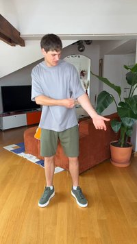 Jeune homme en tenue décontractée retroussant la manche de sa chemise grise dans un salon avec un canapé orange et une plante en pot.