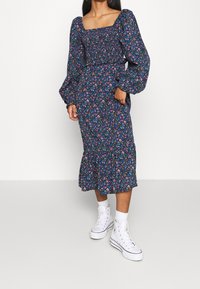 Meklēšana garām piedurknēm, kvadrātveida kakla izgriezums, jūras zils ar ziedu rakstu. Iezīmē puffētas piedurknes, smocked krūšu daļa un pušķota apakša. Valkāta ar baltām sporta kedām.