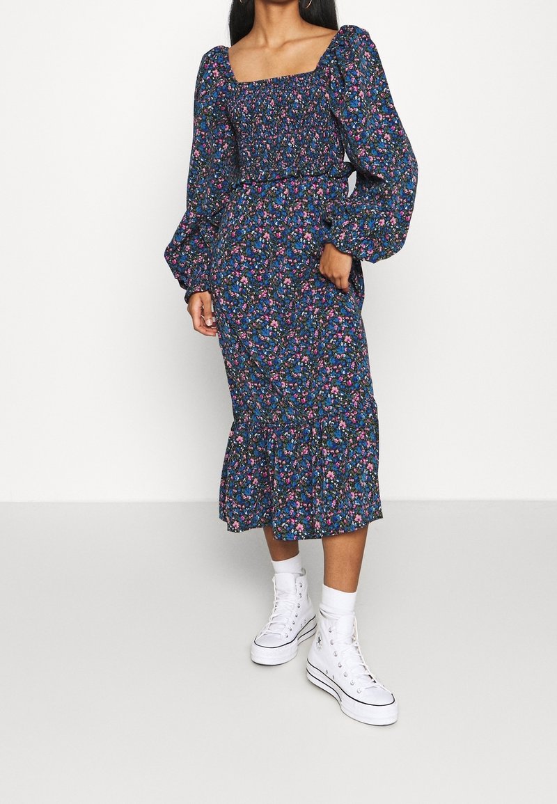 Meklēšana garām piedurknēm, kvadrātveida kakla izgriezums, jūras zils ar ziedu rakstu. Iezīmē puffētas piedurknes, smocked krūšu daļa un pušķota apakša. Valkāta ar baltām sporta kedām.