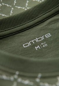 Olivově zelené tričko s žebrovaným výstřihem, se bílou etiketou zobrazující "ombre M eur 48." Tkanina vypadá měkce s texturovaným povrchem.