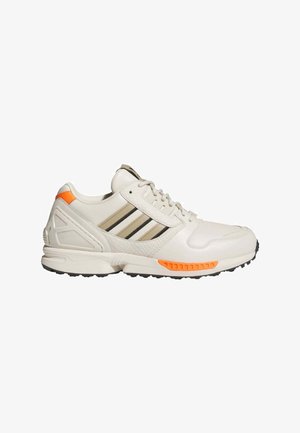 White leather sneaker with black stripes, orange accents, textured heel, and a rugged black sole. Features padded collar and lace-up design.