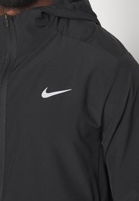 Chaqueta negra de Nike con cremallera frontal, capucha y un logo bordado en blanco en el lado izquierdo del pecho. Material suave y ligero.