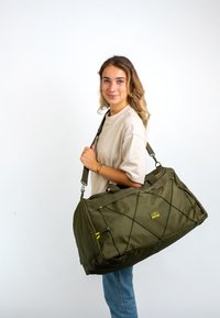 Olive zelená sportovní taška s texturovanou tkaninou, pevnými madly a nastavitelným popruhem. Obsahuje žluté akcenty a křížový design na přední straně.