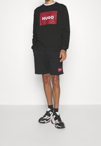 Sudadera negra con un gráfico de logotipo en rojo y blanco; combinada con pantalones cortos negros que tienen un parche de logotipo similar. Llevado con zapatillas negras y blancas.