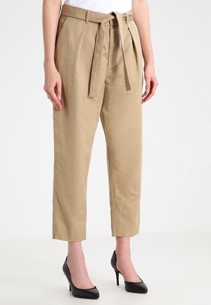 Femme portant un pantalon beige taille haute fuselé avec ceinture en tissu nouée à l'avant, associé à des escarpins noirs pointus, debout sur un sol blanc.