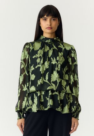Kvinde med langt mørkt hår iført en sort bluse med grønne blomsterbroderier og gennemsigtige lange ærmer, stående mod en ensfarvet baggrund.