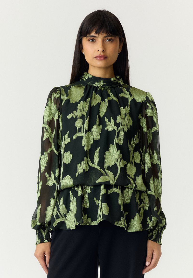Femme aux longs cheveux foncés portant un chemisier noir avec une broderie florale verte et des manches longues transparentes, debout devant un fond uni.