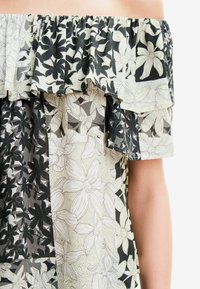 Top off-shoulder con rouches a strati, realizzato in un tessuto floreale bianco e nero a fantasia, con diverse texture e forme.
