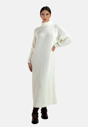 Frau trägt ein langes, weißes, gestricktes Rollkragenpulloverkleid mit Puffärmeln und braunen Stiefeletten, steht vor einem schlichten Hintergrund.