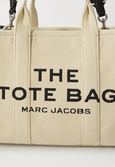 Beżowa torba zakupowa z płótna z czarnym napisem "TORBA MARC JACOBS". Posiada wytrzymałe uchwyty i zamek błyskawiczny. Gładka tekstura.