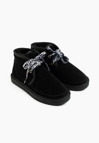 Botas de tobillo de ante negro con punta redondeada, que presentan cordones a rayas de cebra en negro y blanco, ojales dorados y una suela de goma texturizada.