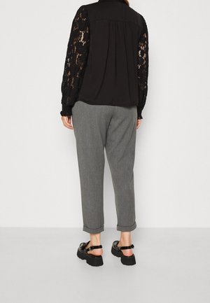 Femme portant un haut en dentelle noire à manches longues, un pantalon gris retroussé et des sandales noires à semelle épaisse, debout devant un fond clair uni.