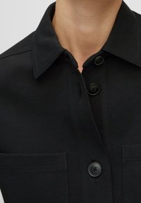 Černá košile s knoflíky, která má límec, dvě přední kapsy a detaily v šití. Tkanina vypadá hladce a má mírný lesk.