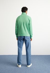 Sweatshirt verde com fecho na frente e gola alta, usado sobre jeans de ganga azuis e sapatilhas brancas, visto de costas. Textura suave.