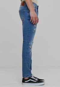 Kék farmernadrág slim fit szabással, elhasználódott részekkel és egy világosbarna bőr címkével a hátsó részén. Fekete sportcipőkkel párosítva.