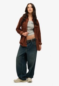 Bruine knoopshirt, grijs getailleerd tanktop, losse donkere spijkerbroek, gestikte riem en lichte sneakers; casual, ontspannen fit.