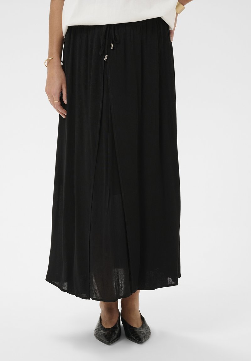 Femme portant une longue jupe noire fendue sur le devant, associée à des chaussures noires à bouts pointus et un haut blanc, debout sur un fond blanc.
