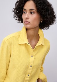 Femme aux cheveux bouclés portant une chemise en velours côtelé jaune, regardant sur le côté contre un fond clair uni.