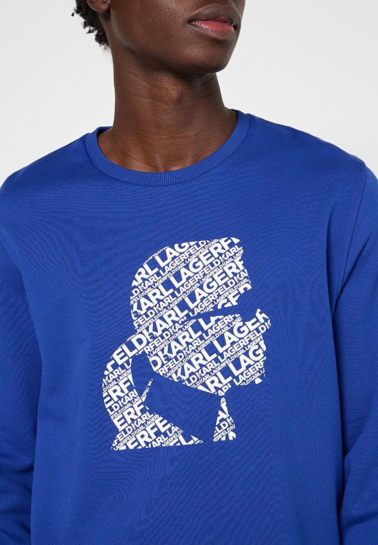 Sweatshirt bleu avec un grand logo graphique blanc composé de texte répété, mettant en avant un tissu lisse et texturé et un col rond.