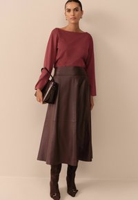 Frau trägt ein burgunderrotes Langarmoberteil, einen dunkelbraunen Leder-Midi-Rock, eine passende Handtasche, goldene Creolen und dunkelbraune Stiefel.