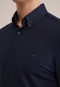 Donkerblauwe polo in een textuurstof. Beschikt over een button-down kraag en een subtiel geborduurd logo op de borst. Twee knopen zichtbaar.