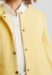 Gelbe strukturierte Jacke mit einer Knopfleiste, die einen Kragen hat und zwei Seitentaschen bietet. Getragen über einem weißen Hemd.