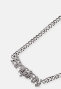 Colar de metal prateado com uma corrente grossa, apresentando um design de placa de nome ousado e estilizado em uma fonte elegante e cursiva.