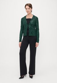 Veste verte à sequins avec capuche, deux boutons à l'avant, associée à un haut noir et un pantalon à rayures noires, le tout complété par des escarpins noirs.