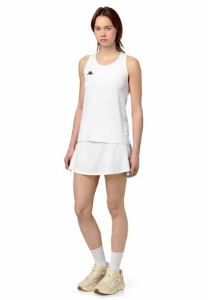 Jeune femme portant un haut blanc sans manches, une jupe blanche, des chaussettes blanches et des chaussures de sport beiges, debout sur un fond blanc.