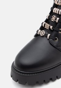 Bottine en cuir noir avec une semelle épaisse à crans, ornée de clips métalliques décoratifs pour les lacets et de strass brillants sur les lacets.