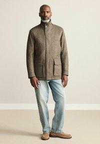 Pruun ruuduline jakk kõrge kaelusega ja kahe esikoti ning helesiniste teksade ja beežide kingadega; sile, tekstuuriga kangas.