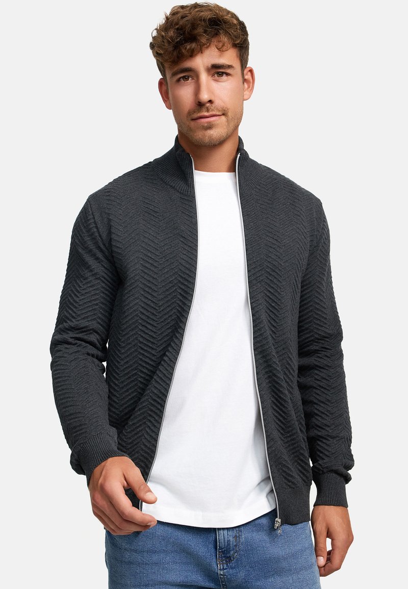 Chaqueta gris con cremallera y patrón en forma de chevrón, con puños y cuello acanalados. Lleva encima una camiseta blanca, combinada con vaqueros azules.