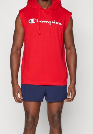 Sudadera sin mangas roja de Champion con logo blanco, combinada con pantalones cortos azul marino, mostrada en una persona de pie desde el cuello hasta la mitad del muslo.