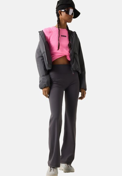 Kobieta w czapce z daszkiem i okularach przeciwsłonecznych, w jaskrawo różowym krótkim topie, szarej puchowej kurtce, dopasowanych szarych spodniach i białych trampkach, stojąca bokiem.