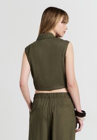 Donna che indossa un top corto senza maniche color verde oliva e pantaloni a vita alta, con le mani in tasca e di schiena rispetto a uno sfondo neutro.