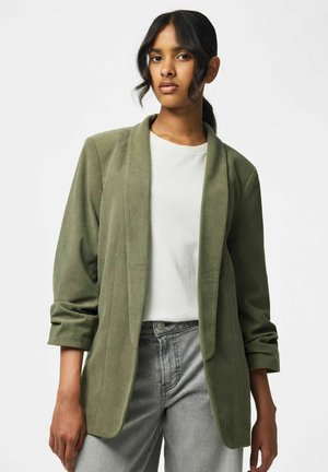 Vrouw met donker haar, gekleed in een olijfgroene corduroy blazer, wit shirt en grijze jeans, staand tegen een egale lichte achtergrond.