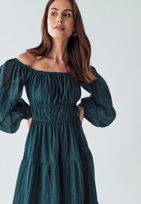 Tirkizna haljina bez naramenica s nabranim rukavima, strukirana i s višeslojnim suknjom. Izrađena od lagane tkanine s teksturiranim završetkom.