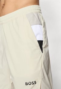 Pantalones cortos deportivos beige con cintura elástica. Presenta paneles de malla en gris, blanco y acentos negros. Logotipo "BOSS" en negro en la pierna.