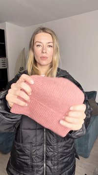 Ribbed pink beanie held in hands, made of soft material, with a fold-over cuff. Background features a teal couch and surfboard.