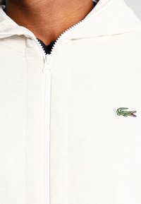Sudadera blanca con cremallera hecha de tela suave, con un logo bordado de cocodrilo verde en el lado izquierdo del pecho y un acabado texturizado.