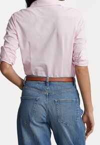 Ljusrosa skjortan med knappar, uppvikta ärmar, i kombination med blå denimjeans och ett brunt läderbälte. Synliga bakfickor på jeansen.