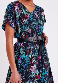 Femme portant une robe noire avec des motifs floraux colorés et de perroquet, un décolleté en V, des manches courtes et une large ceinture noire avec une boucle décorative.
