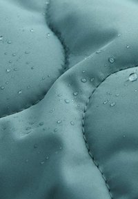 Tissu résistant à l'eau de couleur teal avec une texture lisse, présentant des sections matelassées en relief et des gouttes d'eau à la surface.