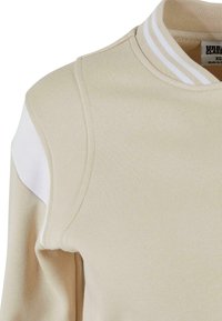 Kortermet beige sweatshirt med ribbestrikket krage. Har en hvit skulderpanel og teksturert stoff. Merket er synlig inne i kragen.