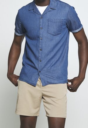 Man wearing short-sleeve blue button-up shirt over white undershirt, paired with beige shorts, standing against plain light background.