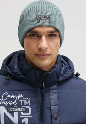 Light blue ribbed beanie with a tag, worn with a dark navy padded jacket featuring zippers and a logo print. Close-up of the upper body.