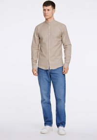 Ανοιχτό καφέ μακρυμάνικο πουκάμισο με γιακά μανδαρινιού, κουμπιά μπροστά, εφαρμοστή γραμμή, σε συνδυασμό με μπλε τζιν και λευκά αθλητικά παπούτσια. Απλός σχεδιασμός, χωρίς σχέδια.