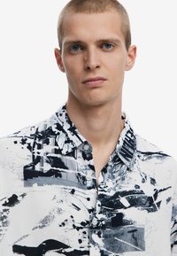 Button-up shirt in white with black abstract patterns, featuring a collar, short sleeves, and a chest pocket with printed text detail.