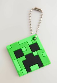 Portachiavi in silicone verde con un design pixelato e squadrato, con accenti neri che formano un volto. Attaccato a una catena in metallo argentato.